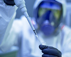 Someone in a laboratory transferring liquid into an epindorf, and wearing PPE including a respirator.