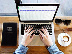 Someone typing on a mac computer, with a journal to the left, data on the screen, and sunglasses and a coffee to the right.